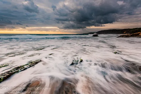 Long-exposure surf over moss-covered rocks at dawn on the New South Wales coast, heavy storm clouds above a warm golden horizon strip.