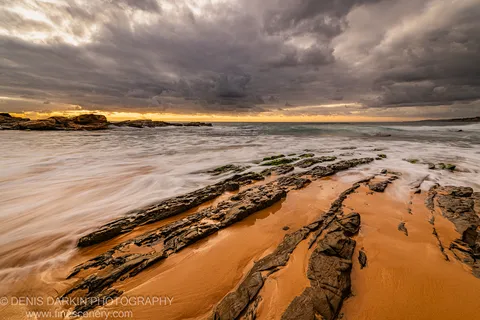 Layered rock with green algae patches extends into milky surf, a warm amber glow at the horizon beneath storm clouds, New South Wales coast.