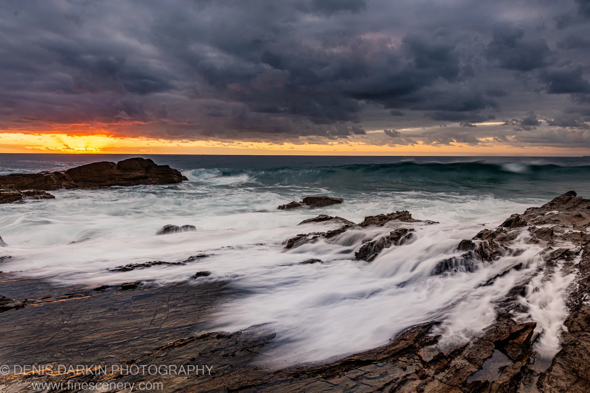 Smooth long-exposure water over layered rock formations on the New South Wales coast, orange sunrise light at the left horizon beneath storm clouds.