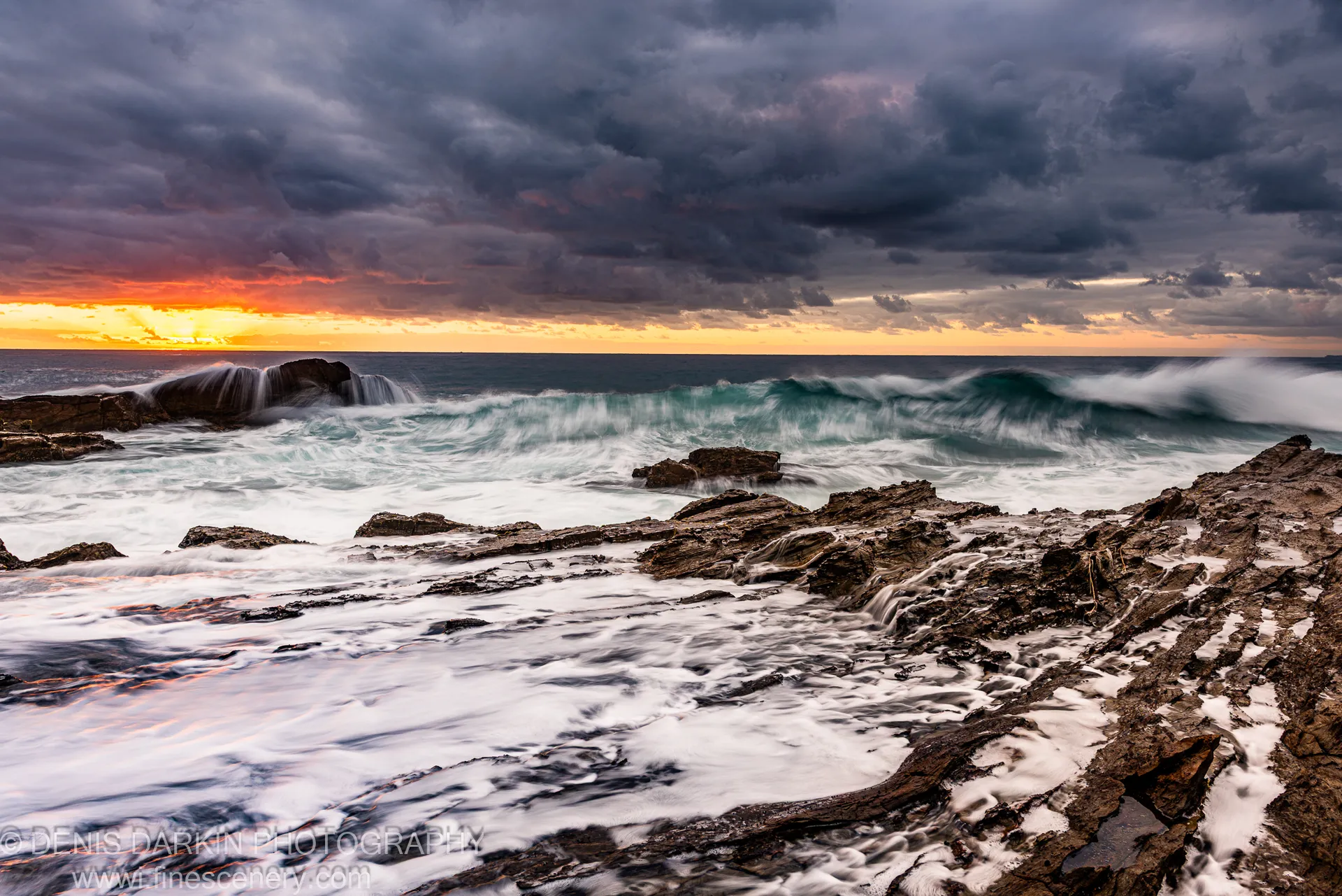 Waves over rugged wet rocks on the New South Wales coast at dawn, an orange sunburst on the horizon beneath dark storm clouds.