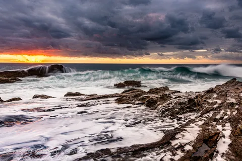 Waves over rugged wet rocks on the New South Wales coast at dawn, an orange sunburst on the horizon beneath dark storm clouds.