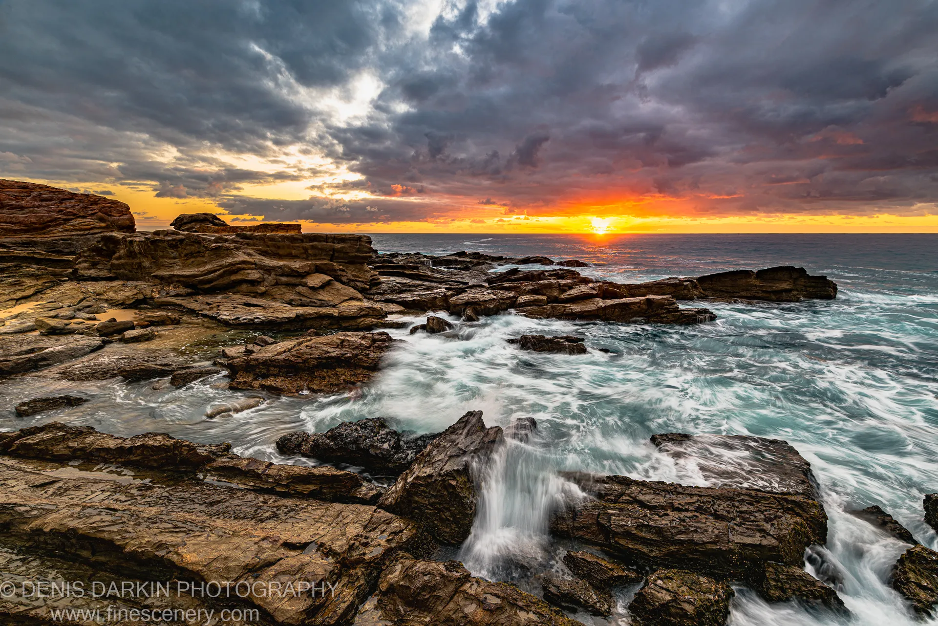 Layered sandstone rock platforms with long-exposure surf in warm orange sunrise light on the New South Wales coast, storm clouds above the horizon.