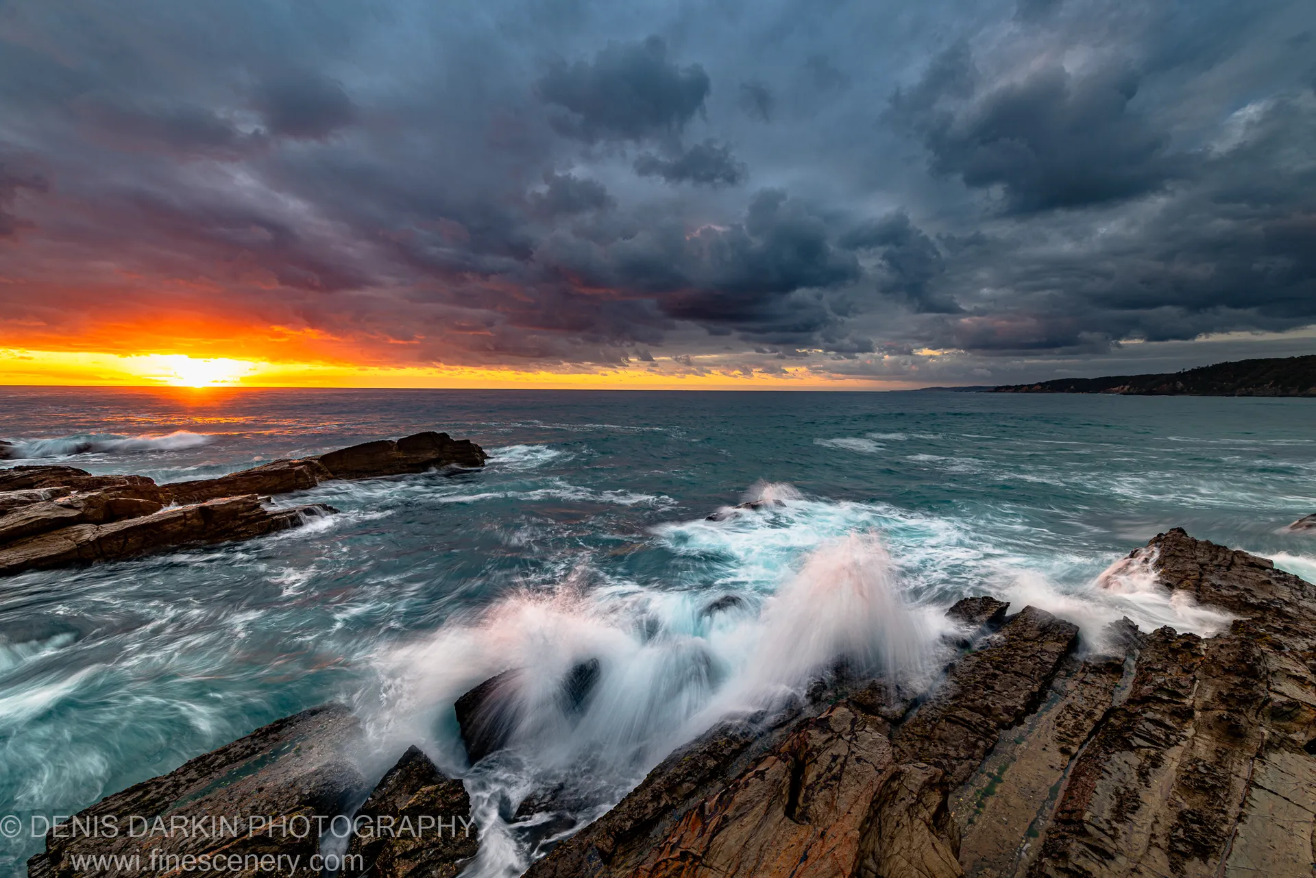White wave spray erupting against layered rock formations at dawn, an orange sunburst through dark storm clouds, New South Wales coast.