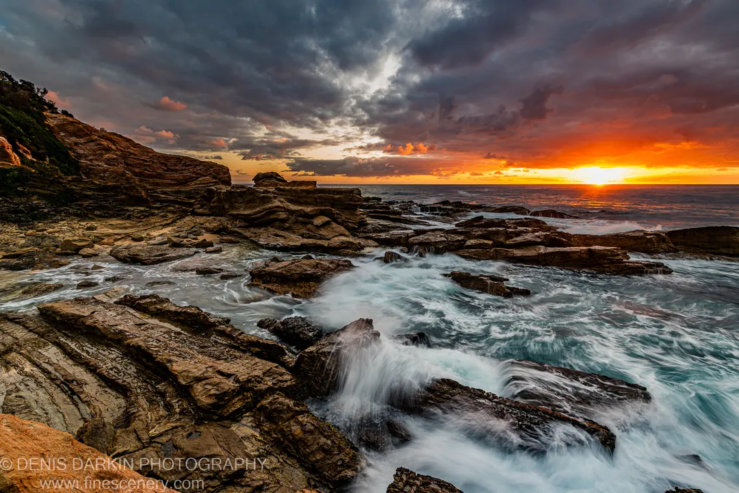 Long-exposure waves flowing through channels in layered rock formations on the New South Wales coast, warm dawn light on the horizon beneath storm clouds.