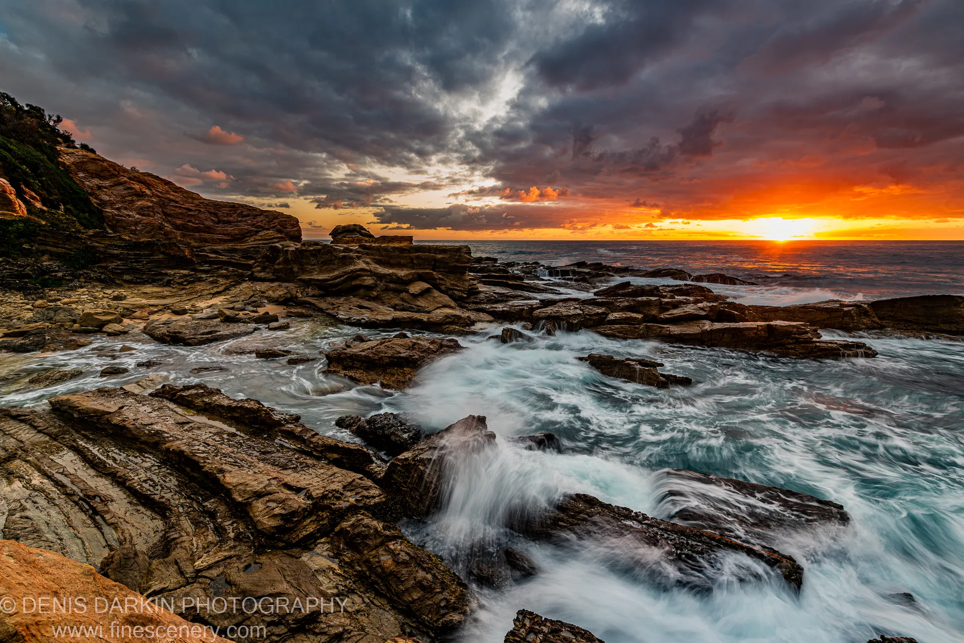 Long-exposure waves flowing through channels in layered rock formations on the New South Wales coast, warm dawn light on the horizon beneath storm clouds.