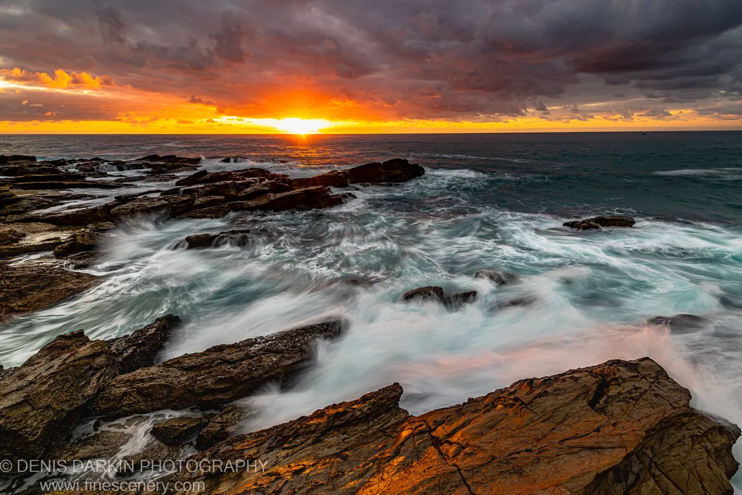Smooth long-exposure water flowing around jagged rock formations on the New South Wales coast at dawn, an orange sunburst emerging from dark storm clouds.
