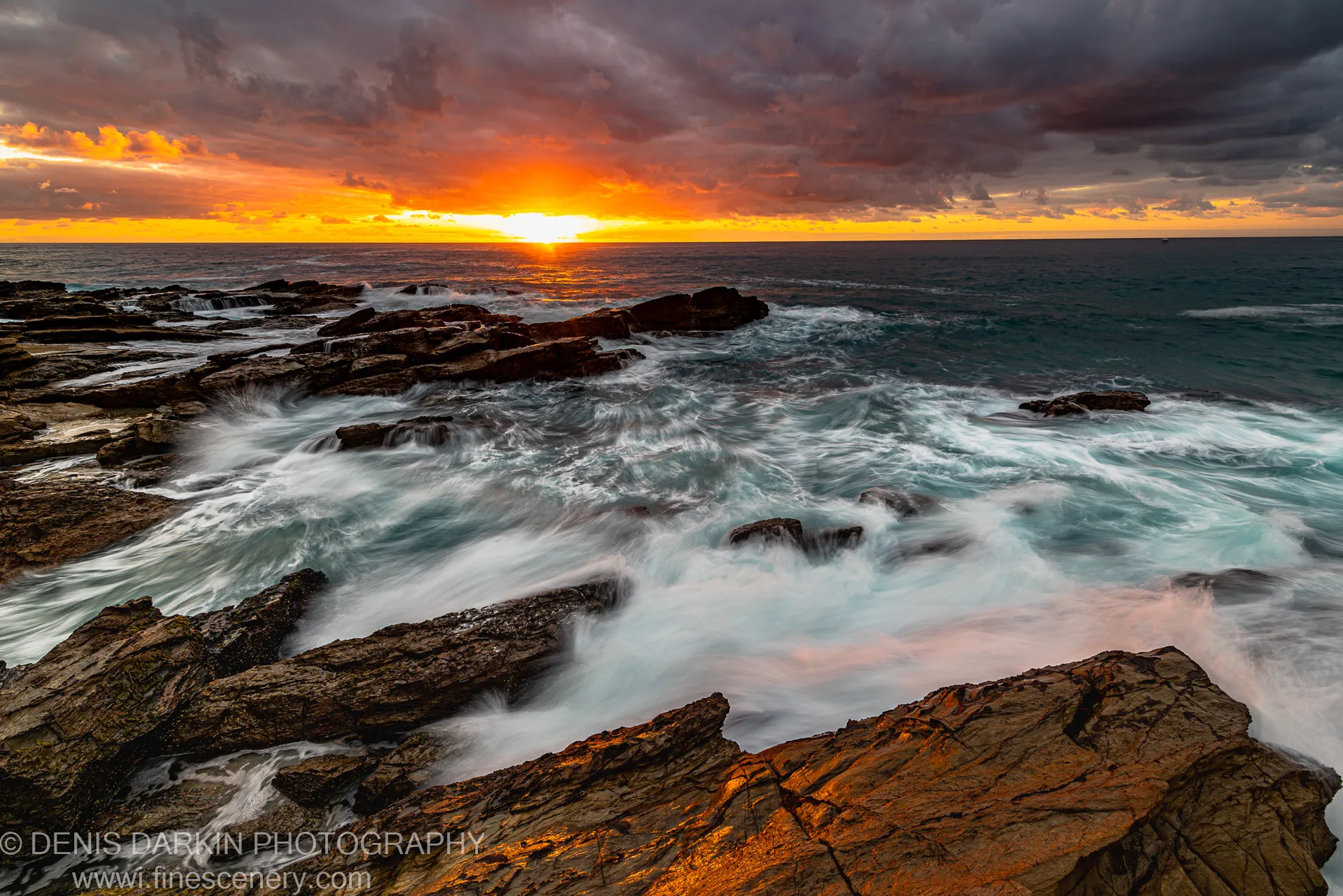 Smooth long-exposure water flowing around jagged rock formations on the New South Wales coast at dawn, an orange sunburst emerging from dark storm clouds.