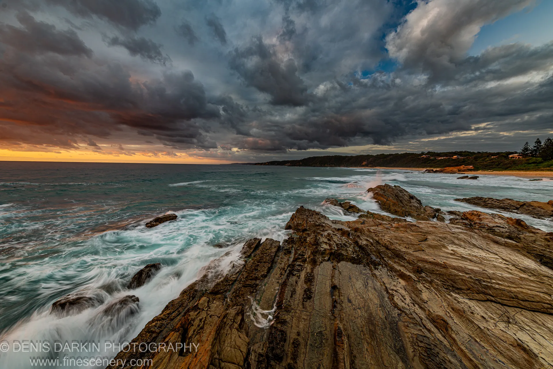Rugged sandstone rocks in the foreground, heavy storm clouds above, and a golden band at the horizon on the New South Wales coast.