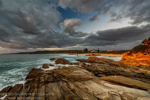 Warm dawn light catches layered sandstone ledges on the New South Wales south coast as storm clouds gather over a secluded sandy bay.