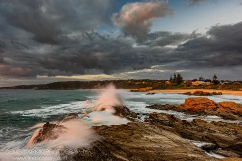 Striated rock formations anchor a view of breaking waves, storm clouds, and distant coastal houses on the New South Wales south coast.