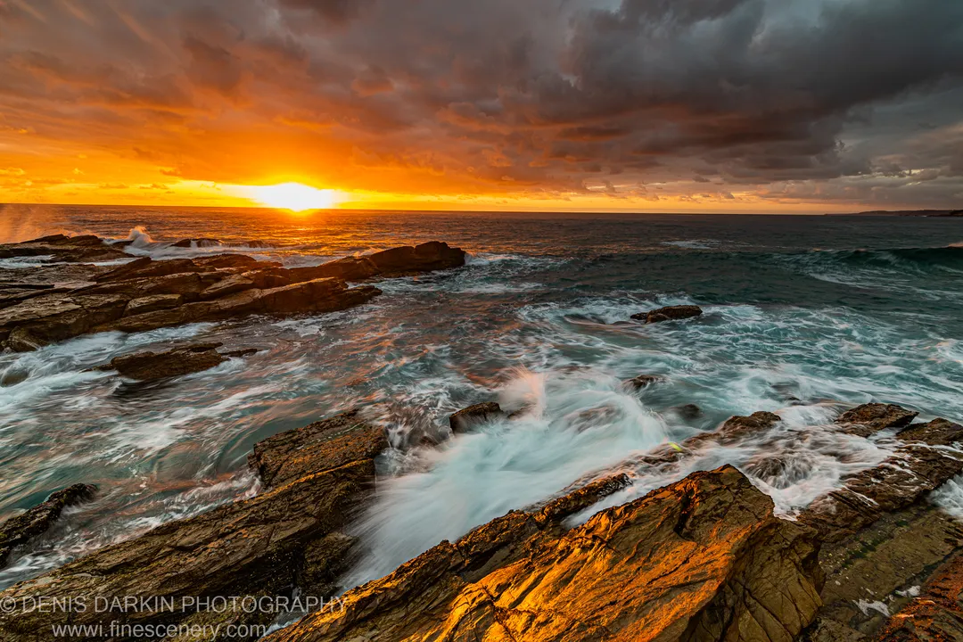 Jagged coastal rocks catch the golden light of a December sunrise while white foam swirls around the dark stone, New South Wales.