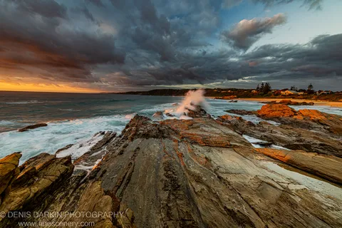 Layered sandstone on the NSW south coast catches amber dawn light as a wave breaks against a central rock, distant houses visible.
