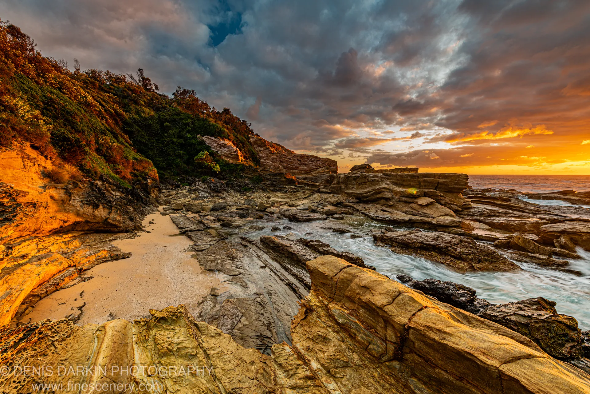 Coastal cliffs in warm directional light overlook a small sandy cove tucked into a rocky inlet, with breaking waves below on the NSW south coast.