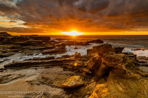 Layered sandstone formations on the NSW south coast catch sunrise light breaking through heavy storm clouds above a turbulent blue-green sea.