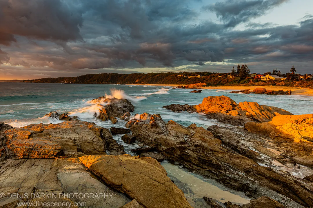 A wave breaks against a dark rock pillar on the NSW south coast at dawn, sending spray into the air above the sunlit stone.
