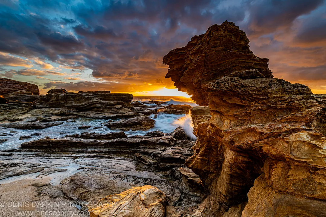 Amber dawn light catches the underside of an overhanging stone pillar on the NSW south coast as long-exposure waves roll across dark, wet shore.