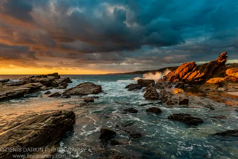 Textured rock strata on the South Coast with calm tide pools reflecting a dual blue and gold dawn sky above the shelf.