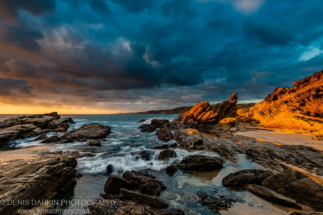 Deep blue storm clouds over a rocky headland at the South Coast NSW at dawn, with warm golden sidelight on the cliff face.