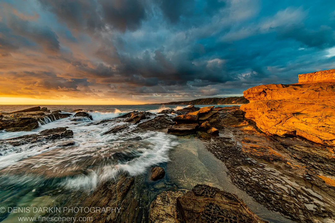 White surf washing over a layered rock platform at dawn on the South Coast, with dark storm clouds above sandstone cliffs behind.