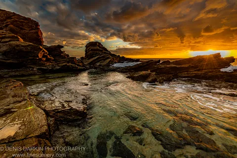 Layered rock pool on the South Coast mirroring an orange sunrise sky, with dark cliff shapes silhouetted against the breaking dawn light.