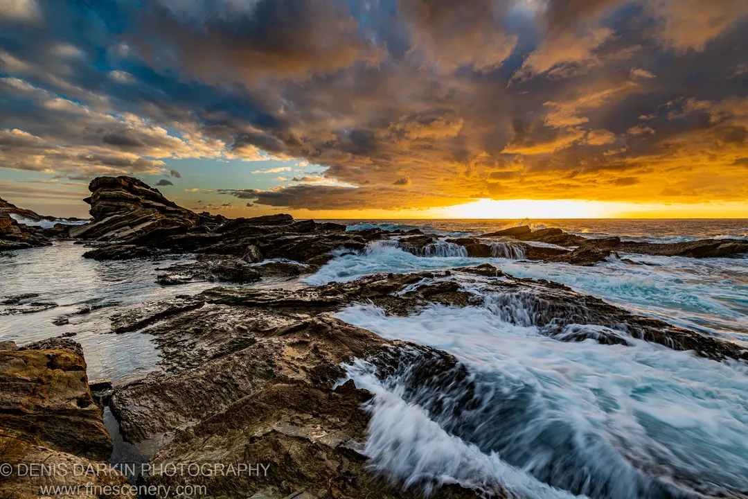 White water over dark layered rock formations at dawn on the South Coast, with a bright horizon band under heavy clouds.