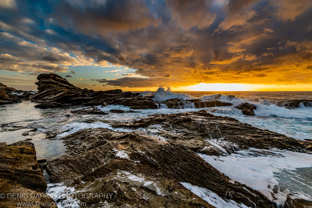 Foamy white waves against dark rock on the South Coast, with deep blue storm clouds and a golden dawn band on the horizon.