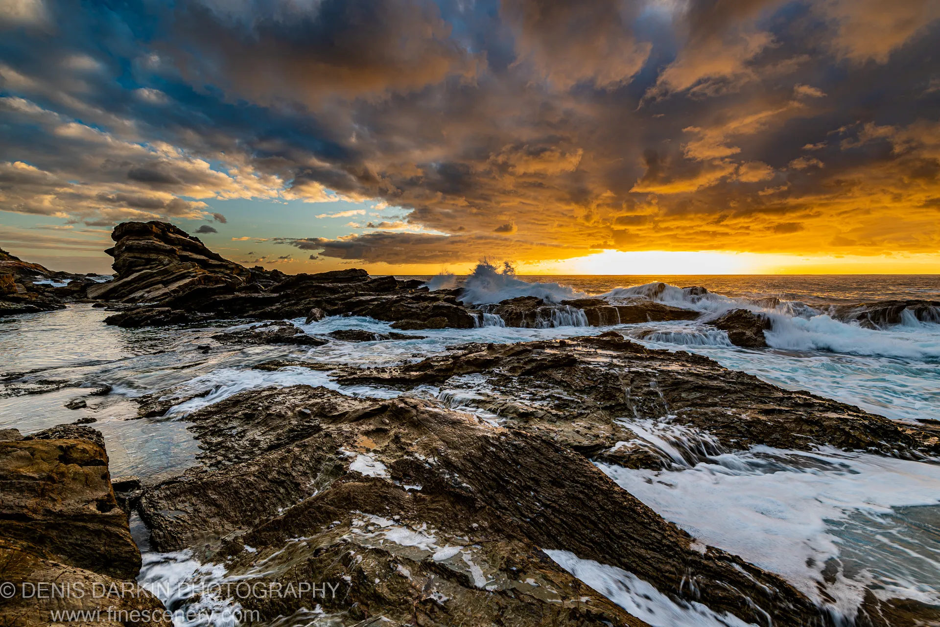 Foamy white waves against dark rock on the South Coast, with deep blue storm clouds and a golden dawn band on the horizon.