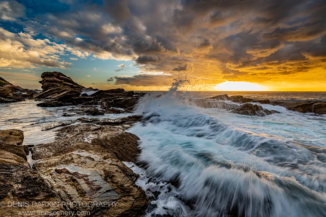 A wave crashing against layered rock at sunrise on the South Coast, with long-exposure foam retreating across the platform.