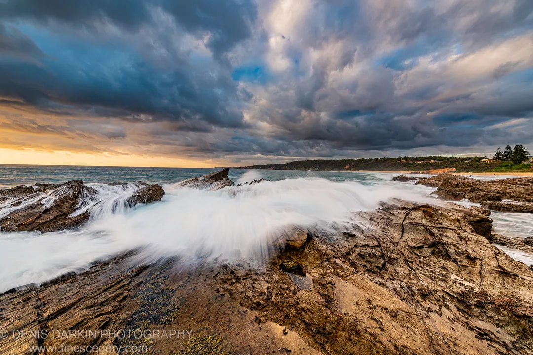 Layered rock formations on the South Coast at dawn, with motion-blurred white water moving over wet stone under heavy grey cloud.