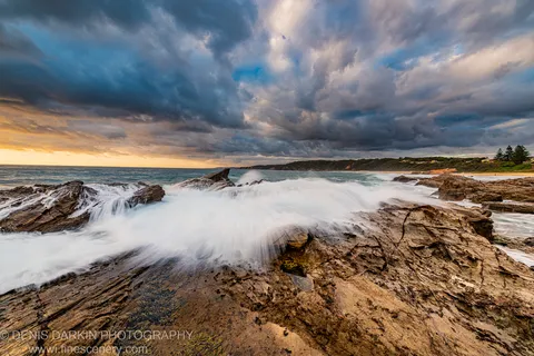 Layered rock formations on the South Coast at dawn, with motion-blurred white water moving over wet stone under heavy grey cloud.