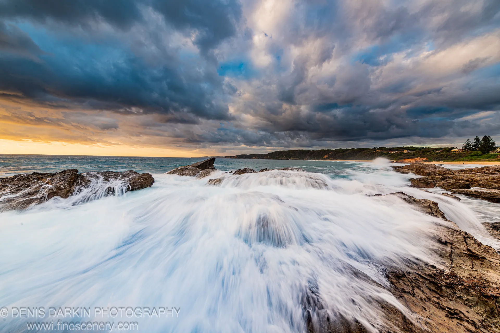 Long-exposure waves flow over dark coastal rocks as storm clouds hang above the New South Wales south coast at dawn.