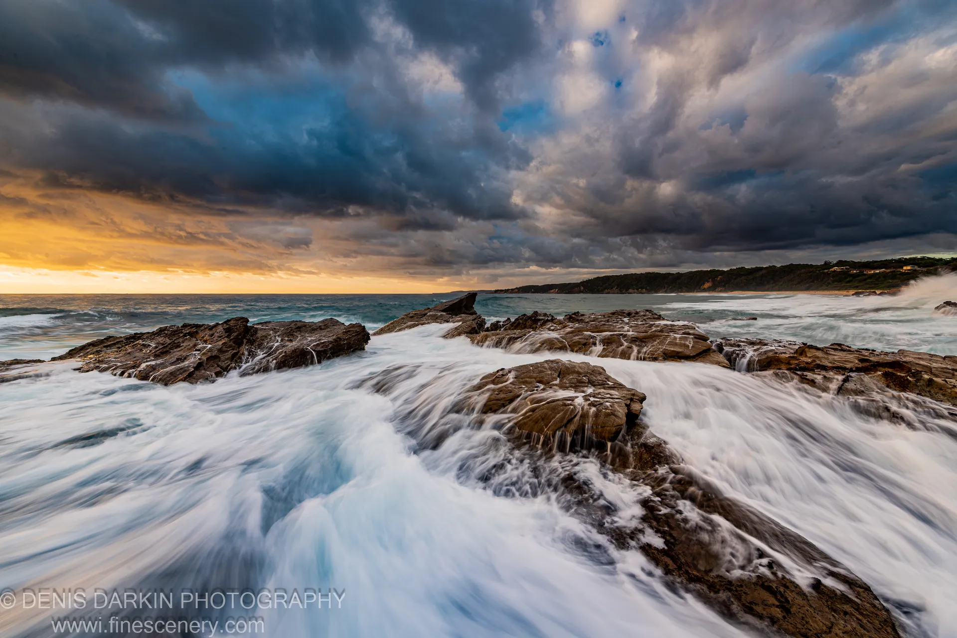 Milky water flows over rugged coastal rocks against a golden dawn horizon and dark storm clouds, New South Wales south coast.