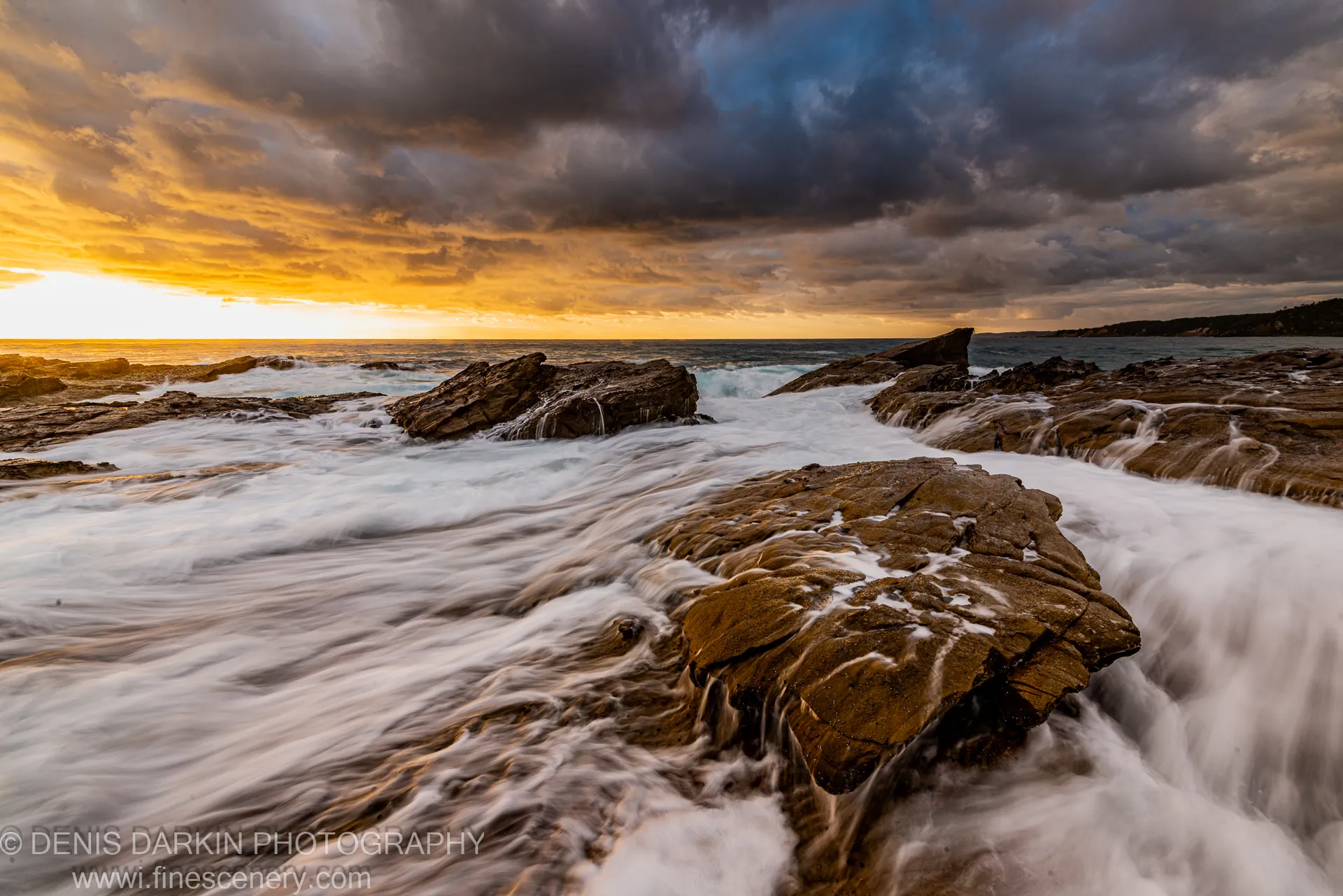 Layered rock formations at the shoreline catch morning light as smooth white water passes over their surfaces, New South Wales south coast.