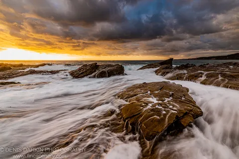 Layered rock formations at the shoreline catch morning light as smooth white water passes over their surfaces, New South Wales south coast.