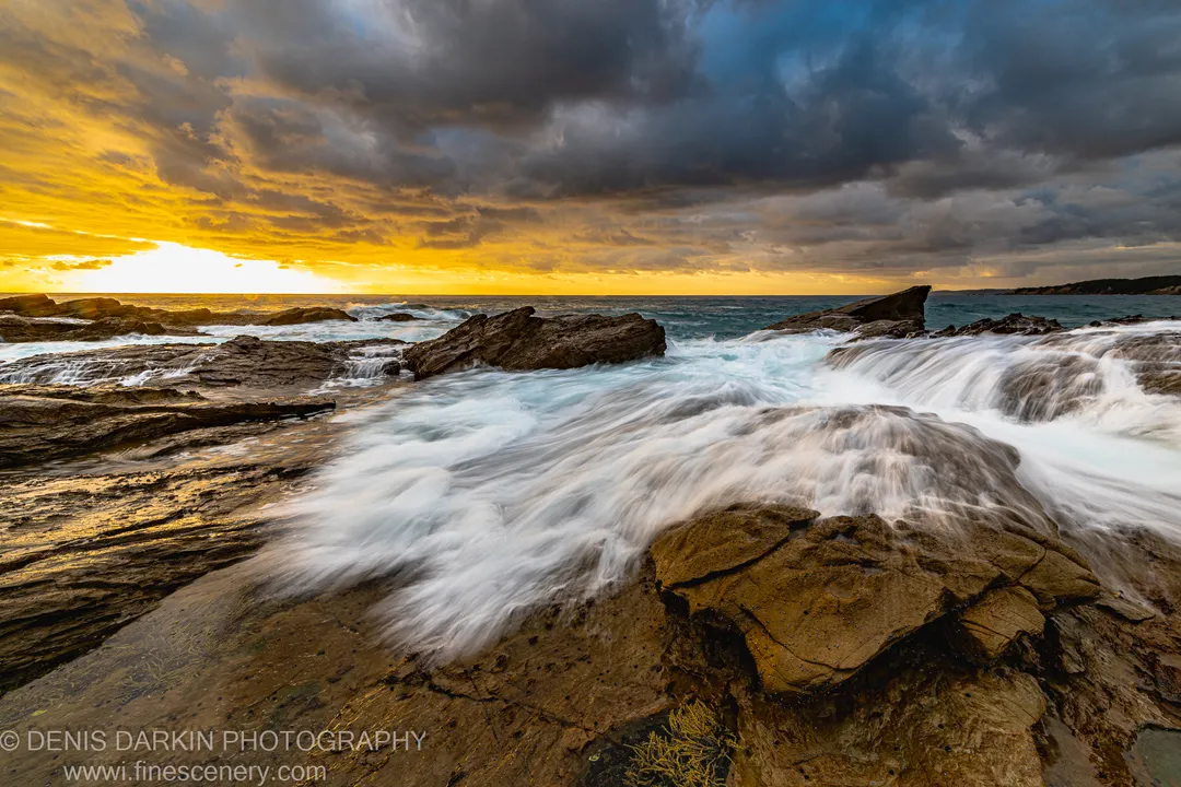White water rushes diagonally across a sandstone rock platform as the sky splits between golden sunrise light and heavy storm clouds.