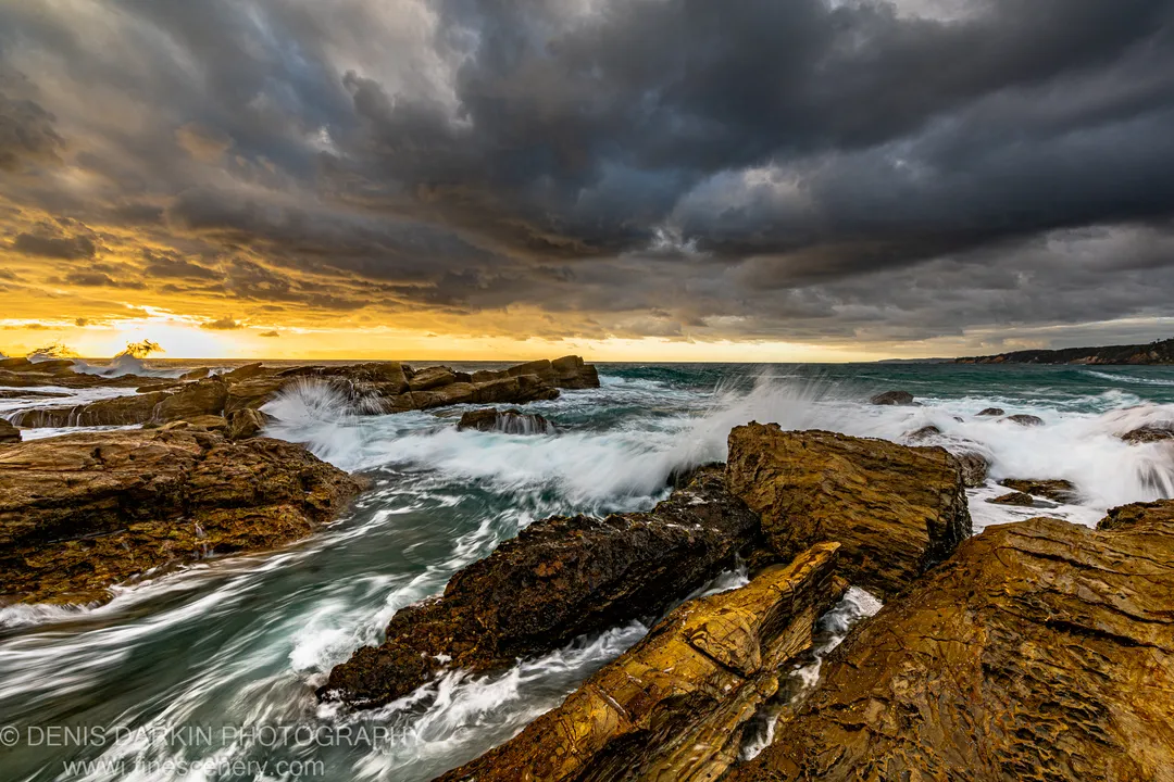Jagged coastal rock formations wet with sea spray catch amber sunrise light as dark storm clouds fill the sky above the New South Wales coast.