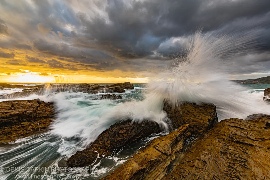 A wave explodes against layered rock formations at dawn, sending white spray skyward while a golden glow breaks through dark storm clouds on the horizon.