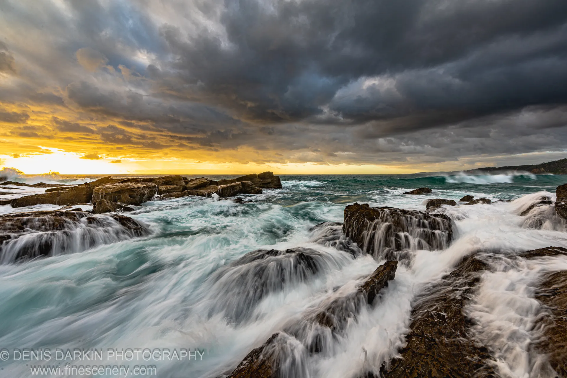 Turbulent waves rush over flat sandstone ledges on the New South Wales south coast at dawn as golden light breaks through heavy storm clouds.
