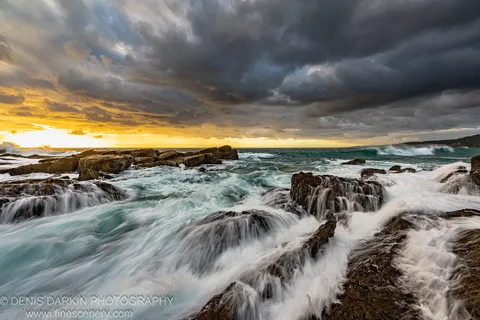 Turbulent waves rush over flat sandstone ledges on the New South Wales south coast at dawn as golden light breaks through heavy storm clouds.