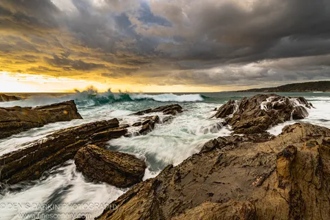 Breaking waves swirl white foam around wet layered rock outcrops as golden sunlight cuts through dark storm clouds above the south coast.