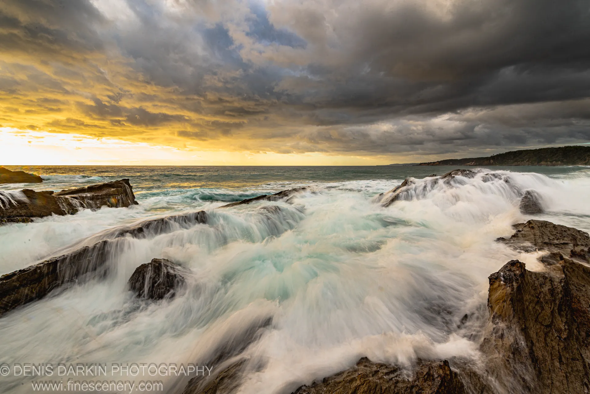 Coastal cliffs and rocky shoreline under a sky divided between golden sunrise light on the left and heavy dark storm clouds on the right.