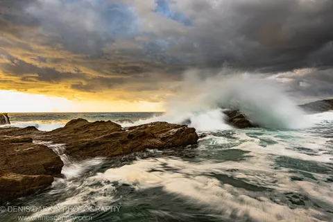 A powerful wave breaks against dark layered coastal rock and sends spray high into the air while smooth water flows over the foreground stone.