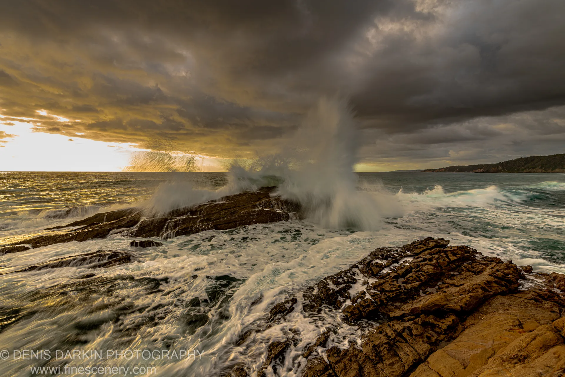 A towering wave breaks against dark wet rocks at dawn on the New South Wales South Coast, spray rising against a golden horizon.