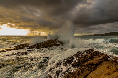 A towering wave breaks against dark wet rocks at dawn on the New South Wales South Coast, spray rising against a golden horizon.