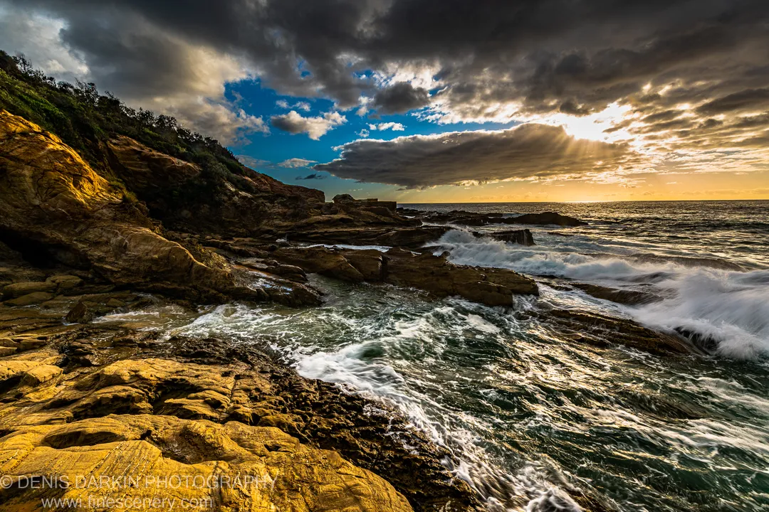 Layered coastal rock catches warm sunlight while waves break below heavy clouds and sunbeams over the New South Wales South Coast.