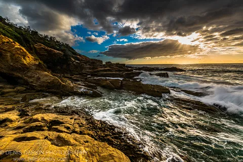 Layered coastal rock catches warm sunlight while waves break below heavy clouds and sunbeams over the New South Wales South Coast.