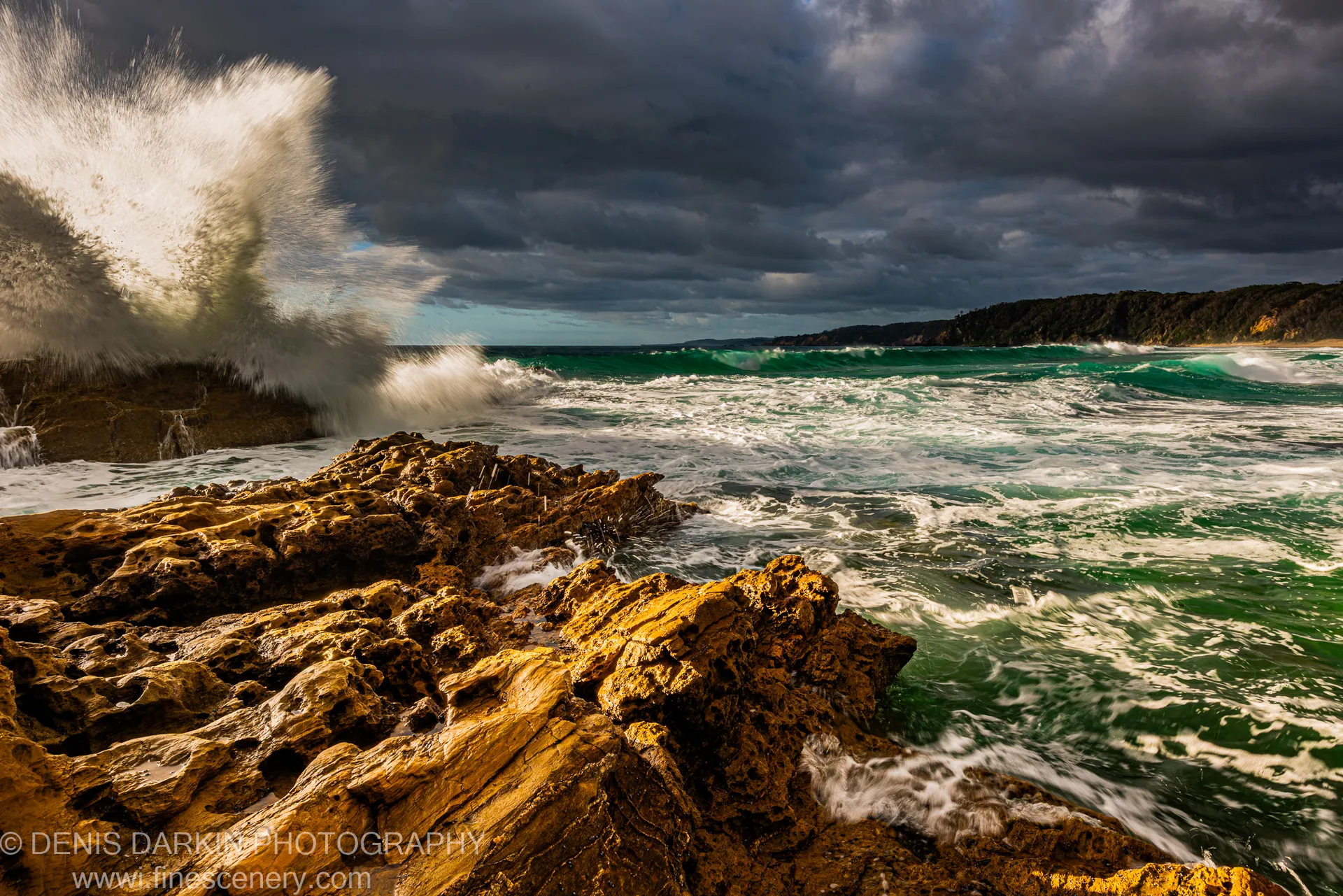 A large wave explodes in white spray against a rocky New South Wales coastline, with a distant vegetated headland and dark storm clouds overhead.