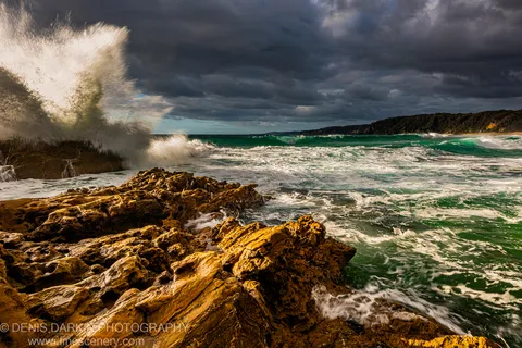 A large wave explodes in white spray against a rocky New South Wales coastline, with a distant vegetated headland and dark storm clouds overhead.