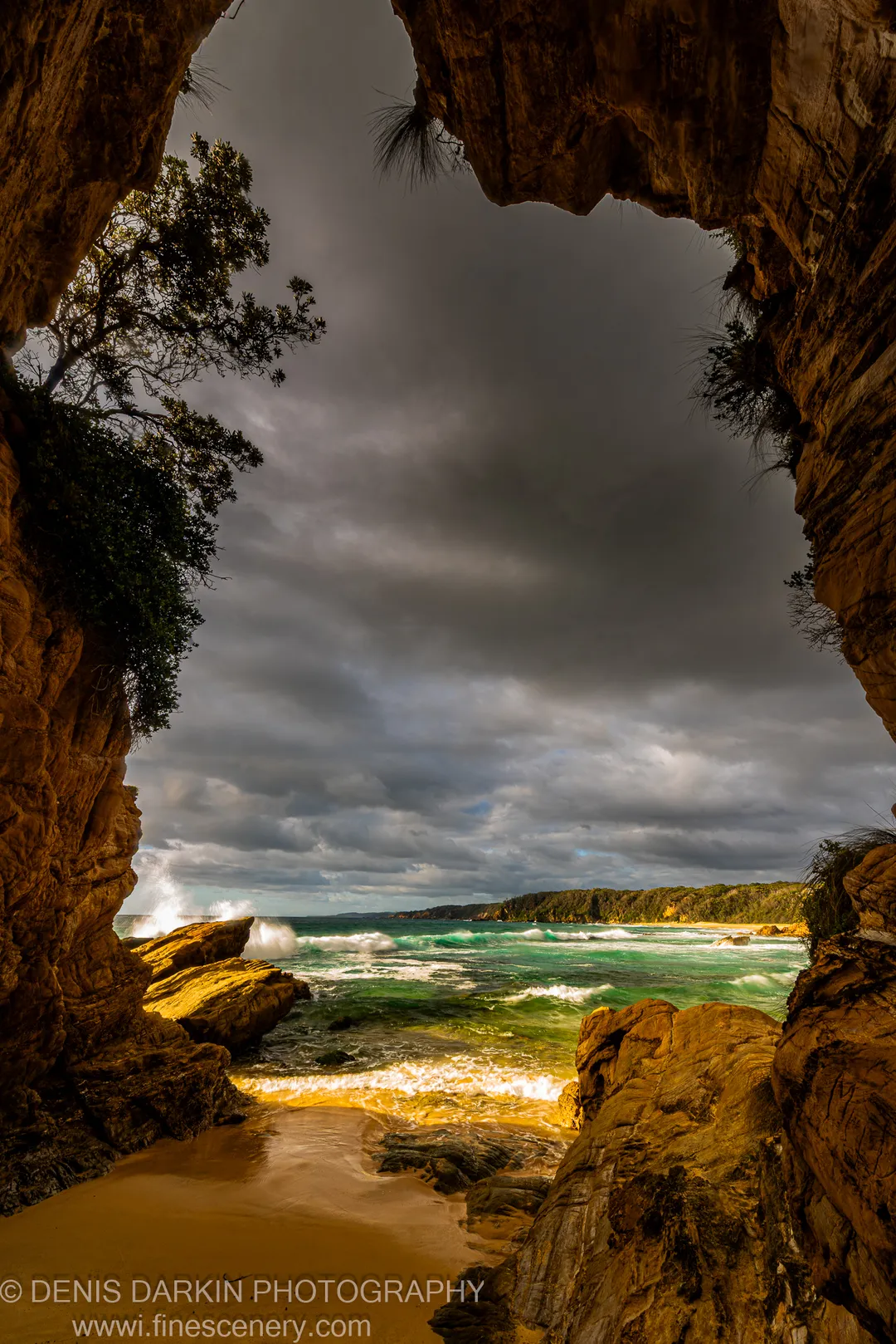 A sandstone archway opens to turquoise surf breaking onto wet sand under storm clouds, South Coast, New South Wales.