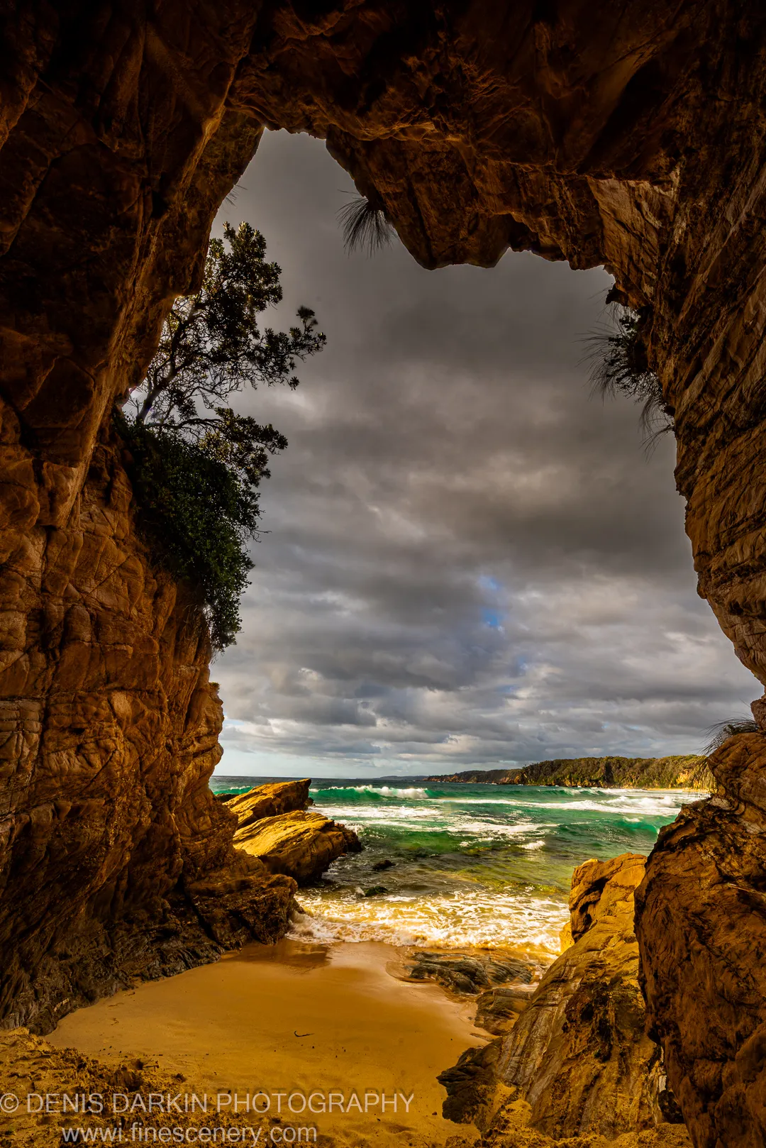Warm amber light fills a sandstone cave opening to turquoise surf and a sandy beach below storm clouds, South Coast, New South Wales.